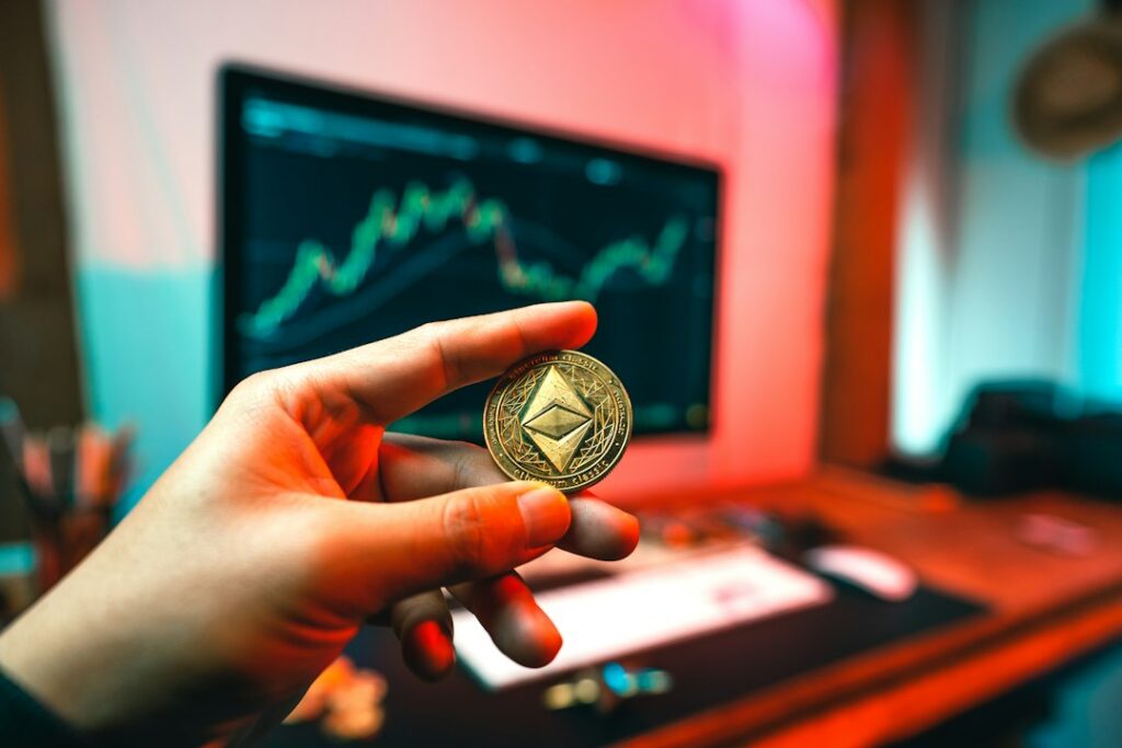 Person holding Ethereum coin in front of computer screen with trading data